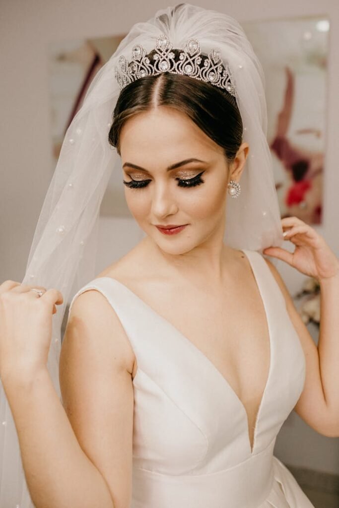 From above of young elegant dreamy woman in white bridal dress with long eyelashes and decorative crown with veil standing with closed eyes